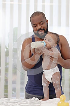 Happy African family, father feeds his toddle baby infant after takes a bath and apply talcum powder on body in bathroom, little photo