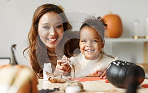 Happy african family mother and cute little girl painting Halloween pumpkins together at home