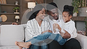 Happy african family parents with cute schoolgirl kid child relax on sofa using funny smartphone app reading joke online