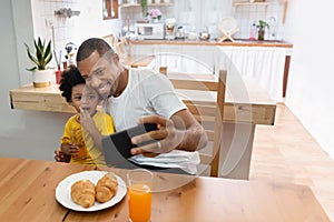 Happy African Father and son taking selfie photo with smartphone during lunch time in dining and kitchen room. Portrait smiling