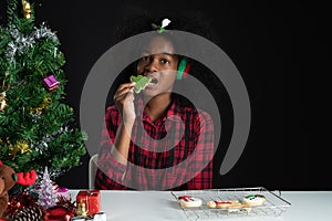 Happy African girl holding and biting Christmas tree shape cookie in hand near little Christmas tree on black background