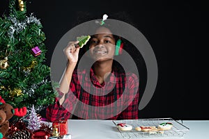 Happy African girl smile and holding Christmas tree shape cookie in hand near little Christmas tree on black background