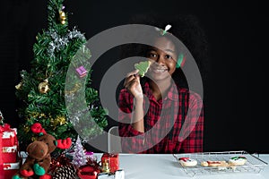 Happy African girl smile and holding Christmas tree shape cookie in hand near little Christmas tree on black background