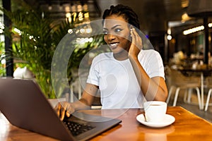 Happy african girl in wireless headphones studying online, using laptop and taking notes in cafe