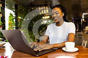 Happy african girl in wireless headphones studying online, using laptop and taking notes in cafe