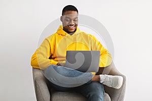 Happy African Guy Using Laptop Sitting In Chair, Gray Background