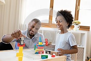 Happy African little son and daddy playing at childish desk