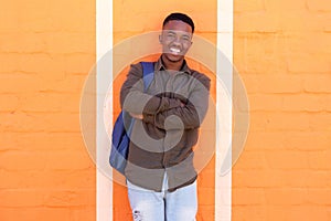 Happy african male student standing against orange wall with bag