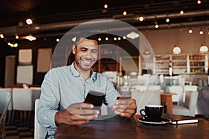 Happy african man holding mobile phone typing card data to make online payment sitting in cafe