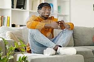 Happy african man using phone while sitting on sofa at his home. Concept of young people working on mobile devices.