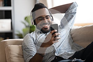 Happy african man wearing headphones listening mobile music on couch