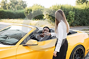 African man sitting inside his new luxury yellow cabriolet car while taking keys from female dealer photo