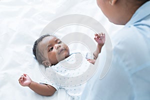 Happy African Nigerian newborn baby lying on white bed at home with mother talking and playing. Innocence infant with curly hair