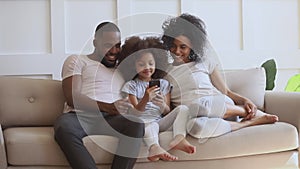 Happy african parents with kid daughter using smartphone on sofa