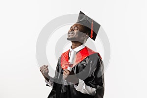 Happy african student in black graduation gown and cap raises masters degree diploma above head on white background