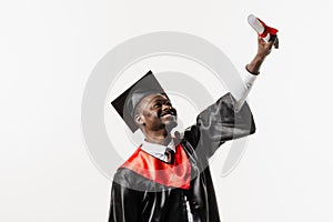 Happy african student in black graduation gown and cap raises masters degree diploma above head on white background