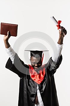 Happy african student in black graduation gown and cap raises masters degree diploma above head on white background