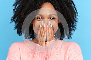 Happy African woman with curly hair smiling covering her mouth and lips with hands, blue background.