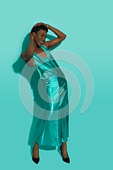 Happy african woman in a turquoise satin long dress is posing with hands on head