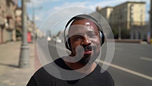 Happy afro-american man is walking on city streets, listening to music by headphones and dancing