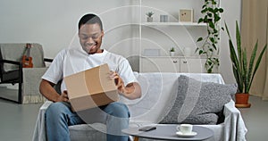 Happy afro man customer open cardboard box receive gift online purchase in postal parcel sittibg on sofa, satisfied