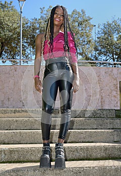 Happy afro-style black girl with long black and blondes braids wearing big shoes, leather pants and sunglasses is standing on