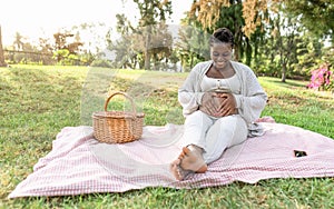 Happy Afro woman touching her pregnant belly doing heart shape with hands in public park