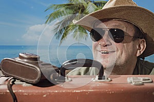 Happy aged man in sunglasses and hat with suitcase and photo camera on tropical beach next to sea, palm tree and blue sky