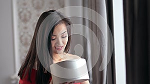 Happy amazed elegant Asian woman opening gift box and smiling at home cozy interior