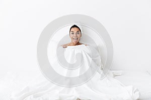 Happy amazing woman with towel on head lies in bed covering herself under blanket isolated over white wall background