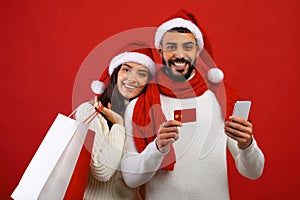 Happy arab couple in Santa hats holding shopping bags, smartphone and credit card, recommending online shopping