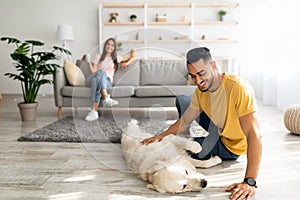 Happy Arab guy stroking cute dog on floor at home, his girlfriend relaxing with cup of coffee on background, copy space