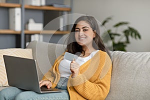 Happy arab woman with laptop and credit card shopping online, using web banking service, sitting on sofa at home