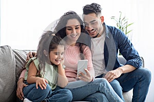 Happy arabic family of three using smartphone at home, relaxing on couch