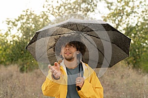 Happy arabic young man with umbrella under the autumn shower. Guy enjoying rainfall on the nature outdoors. Walk in the