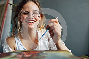 Happy artist female standing next to the easel with canvas painting something in her art studio. A young woman painter in
