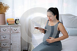 Happy Asia pregnant woman reading book and sitting on white bed
