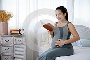 Happy Asia pregnant woman reading book and sitting on white bed