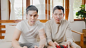 Happy asia young gay couple sit on couch use joystick controller play video game spend fun time together in living room at house.