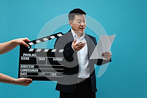 Happy asian actor performing role while second assistant camera holding clapperboard on light blue background, selective focus.