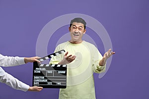 Happy asian actor performing while second assistant camera holding clapperboard on purple background. Film industry