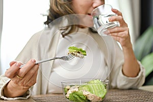 Happy Asian-aged eating salad bowl and drinking water in her dining room. cropped