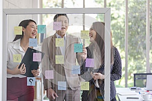 Happy Asian business team meeting planning new strategy using colored sticky notes on mirror board in workplace.