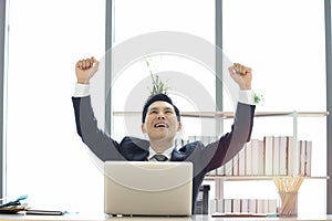 Happy Asian businessman in suit setting at working desk in the office,  raising his arm up to celebrate success goal, man in