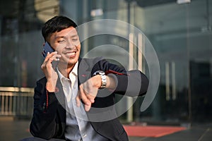 Happy Asian businessman talking on the phone and checking the time on his watch