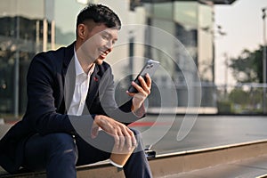 Happy Asian businessman using his smartphone while sitting on stairs outside of the building