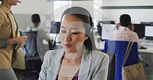 Happy asian businesswoman sitting at desk using computer and talking on headset