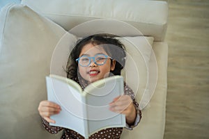 Happy asian child baby girl smiling wearing glasses and reading book while sitting on couch sofa in living room at home. Girl