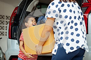 Happy asian child girl and mother helping to carry a cardboard box with stuffs moving into the car together to relocation