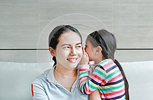 Happy Asian child girl sharing a secret to her mom in the living room at home. Kid girl whispering gossip a something to mother